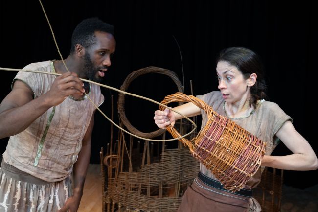 A woman is holding a basket and a man is holding a bent stick. They are both looking at the basket with a shocked expression.
