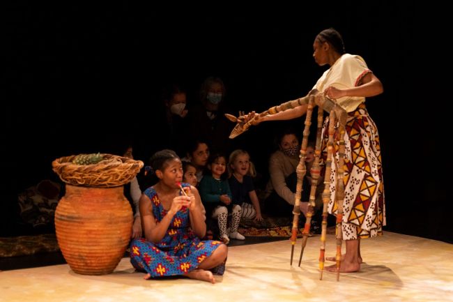 A girl holding a toy snake in the direction of a girl sitting on the floor playing an instrument.