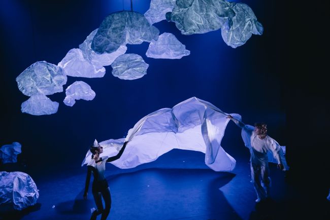 Two people holding a white cloth in the air against a blue background. There are paper clouds at the top of the image.