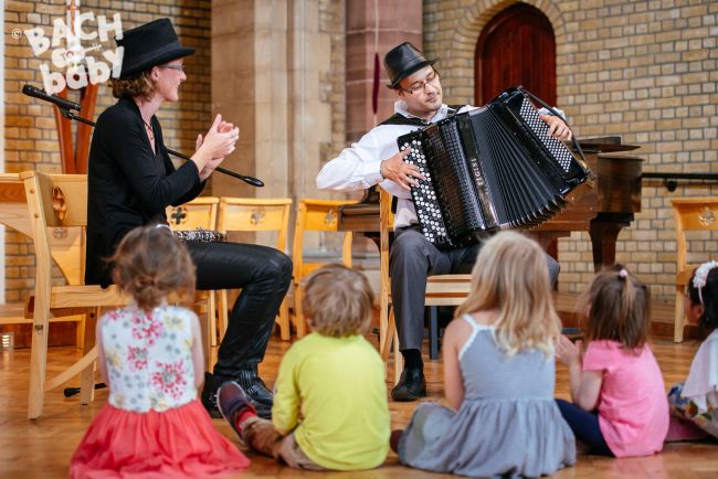 Two musicians wearing black top hats. The lady on the left is clapping and the man on the right is playing the accordion. A row of young children in colourful clothes sitting on the floor in front